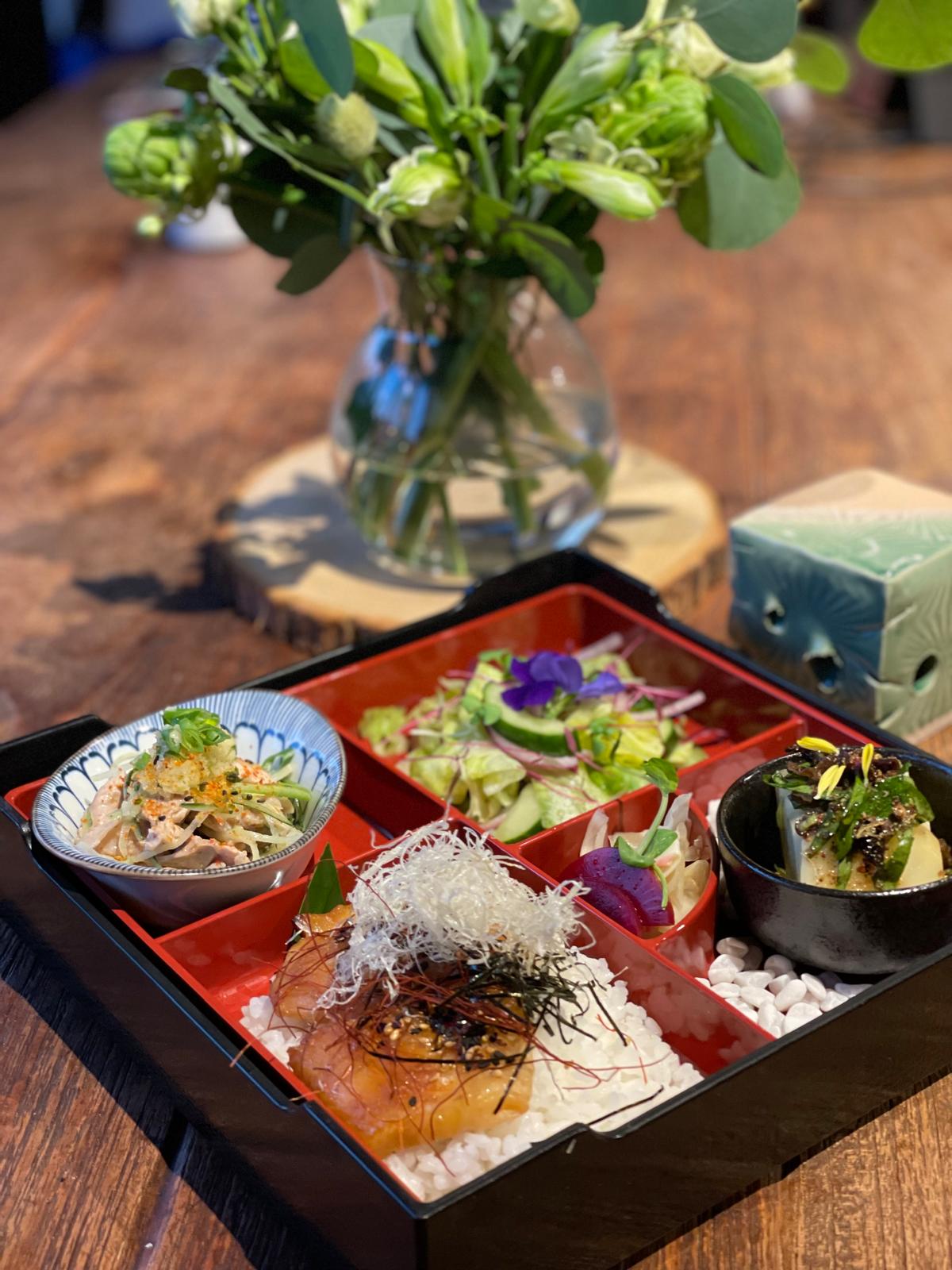 Japanese bento box with various dishes on a wooden table, accompanied by a vase of flowers.