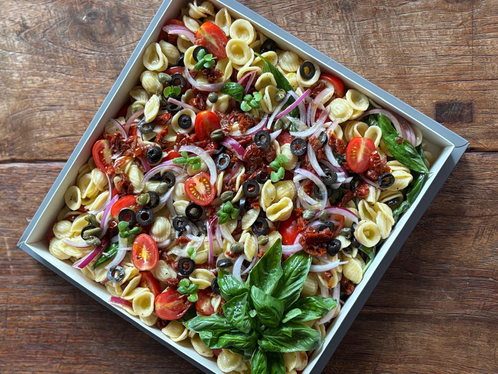 Pasta salad with tomatoes, olives, and basil in a white dish on a wooden table