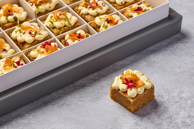 Delicate carrot cake with floral garnish on a gray surface, next to a tray of similar desserts.