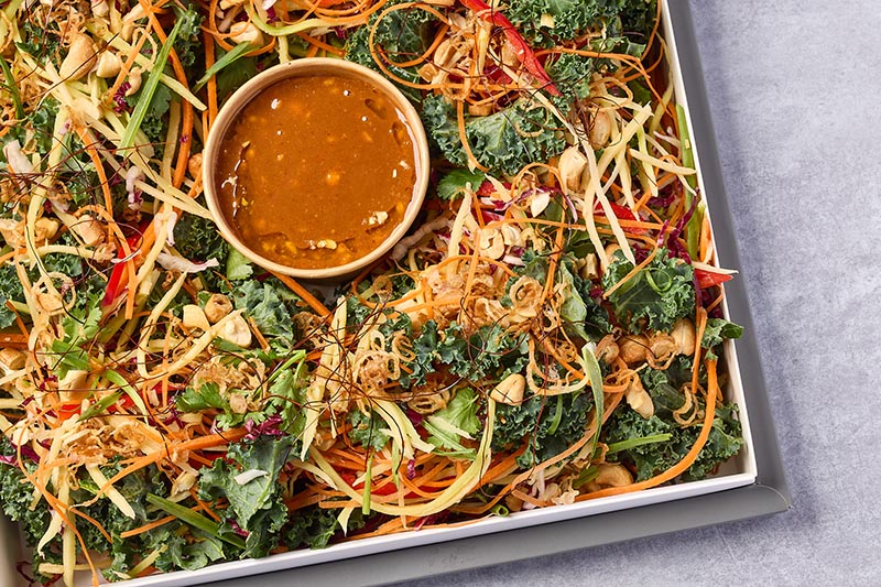 Vegetable salad with kale, carrots, and a small bowl of dressing on a gray surface.
